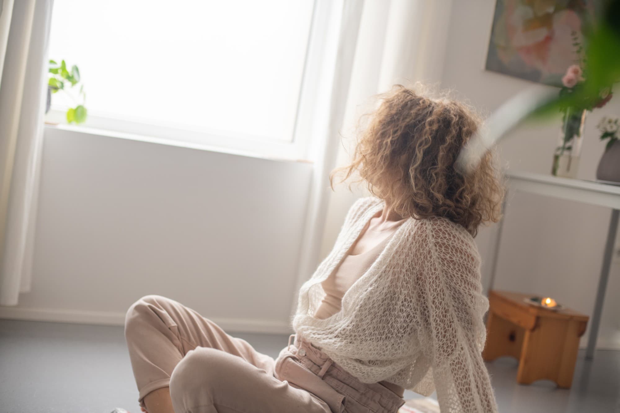 A woman sitting on the floor by a bright window in a cream knit cardigan